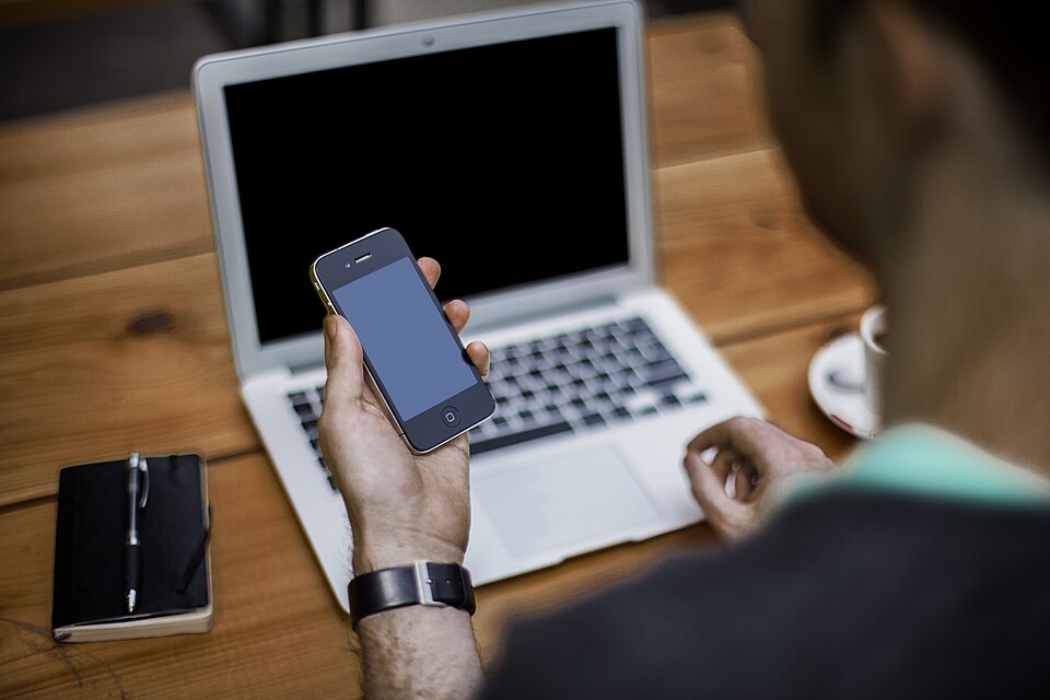 A person holding a phone beside an open laptop on a wooden table.