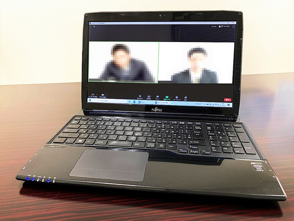 A laptop on a wooden table displaying a video call with two pixelated participants.