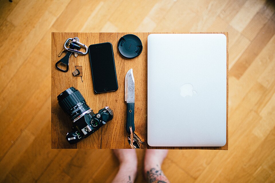 A camera, closed laptop, phone, keys and a small knife arranged on a wooden tabletop.